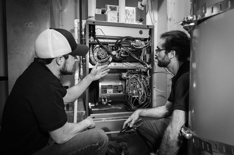Two men examining a furnace in a utility room; one points to components.