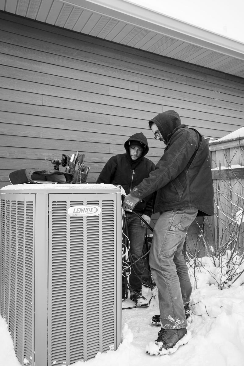 Two people working on an air conditioning unit outside a house in the snow.