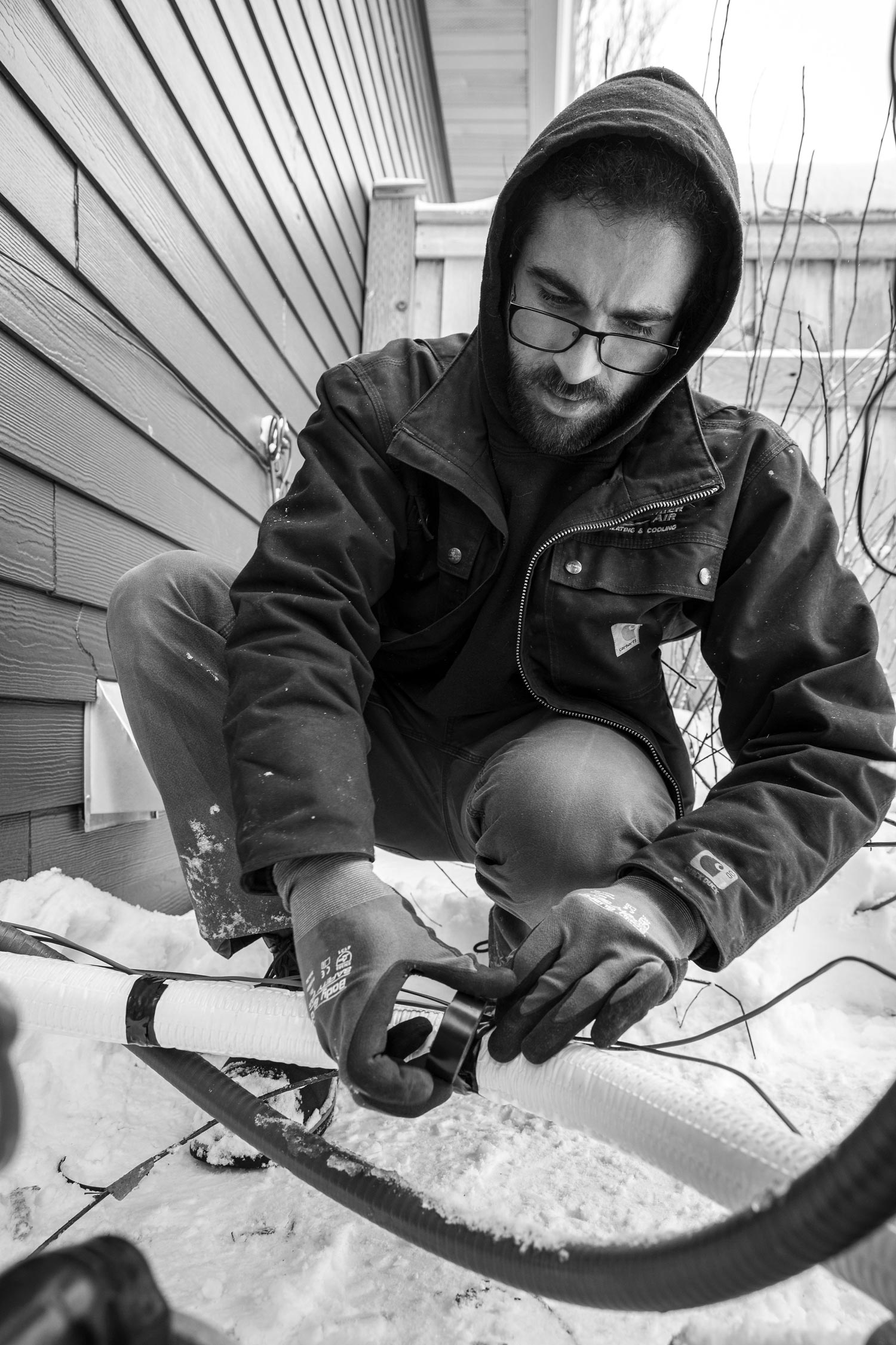 Person kneels in snow, working on a pipe. Wearing hooded jacket and glasses. Exterior setting.