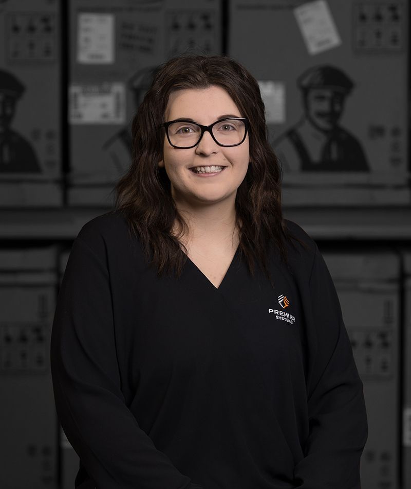 Woman with glasses smiles, wearing black scrubs, standing in a warehouse setting.