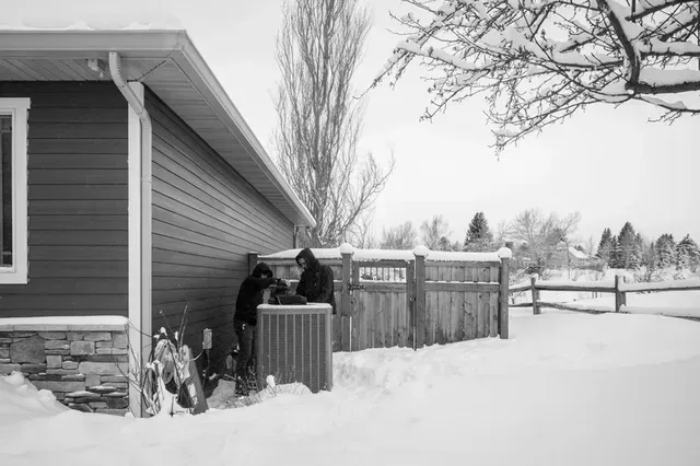 Two people near a snow-covered air conditioning unit next to a house. One is working on it. Winter day.