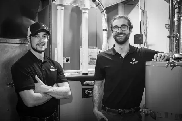 Two men in black shirts smile next to HVAC equipment. One wears a hat, arms crossed.