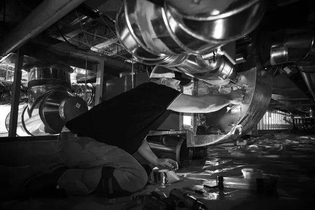 Person kneeling under ventilation ducts, working on equipment. Indoor setting, black and white.
