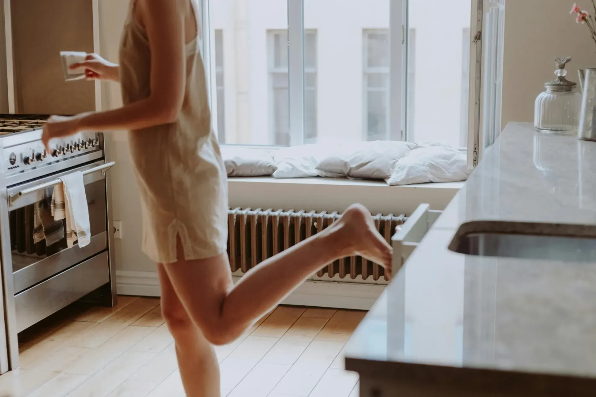 Woman in short beige dress in kitchen, moving forward. Sunlight from window.