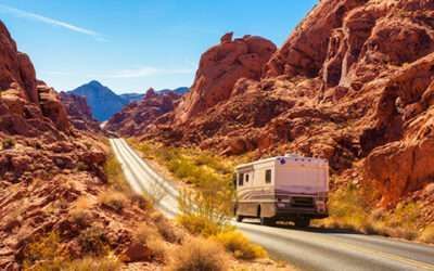RV driving on a winding desert road between red rock canyon walls under a bright blue sky