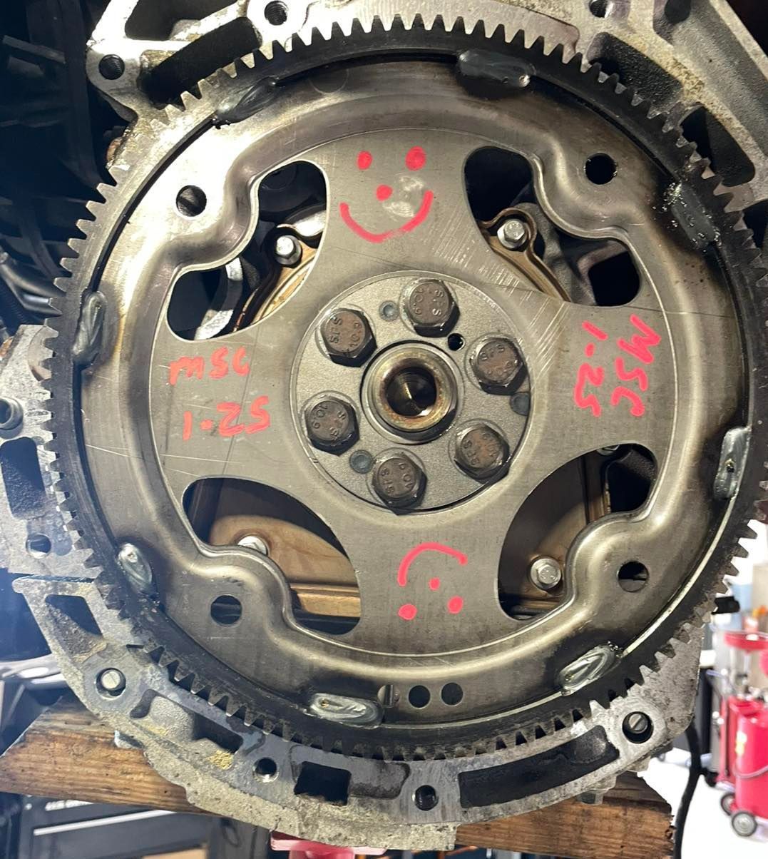 Close-up of a metal flywheel with a gear ring. Red smiley faces are drawn on it.