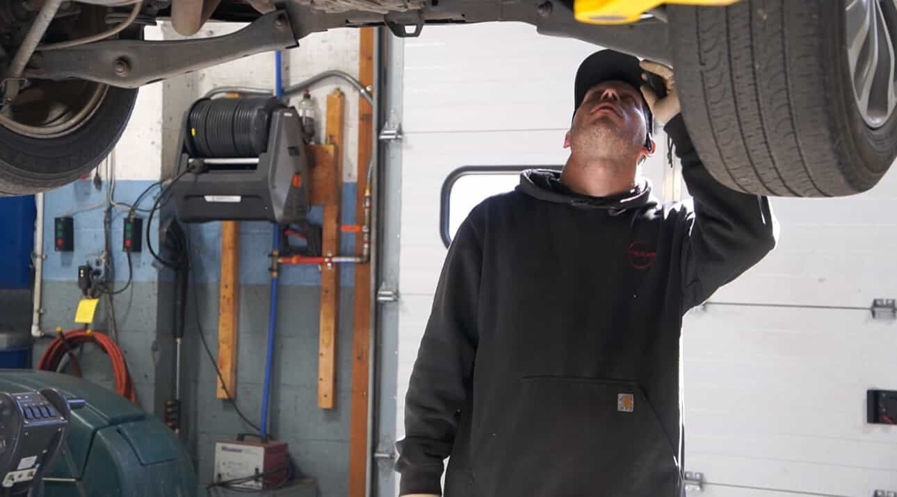 Mechanic in a garage, inspecting the underside of a car lifted on a hoist.