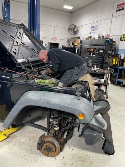 Mechanic working on the engine of a black Jeep with the hood open in an auto repair shop.