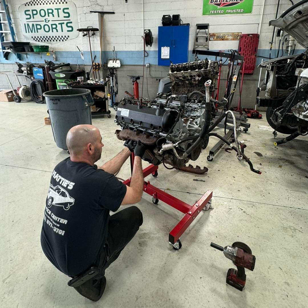 Man working on an engine in a garage. He’s kneeling, using a stand. There's a power tool on the floor.