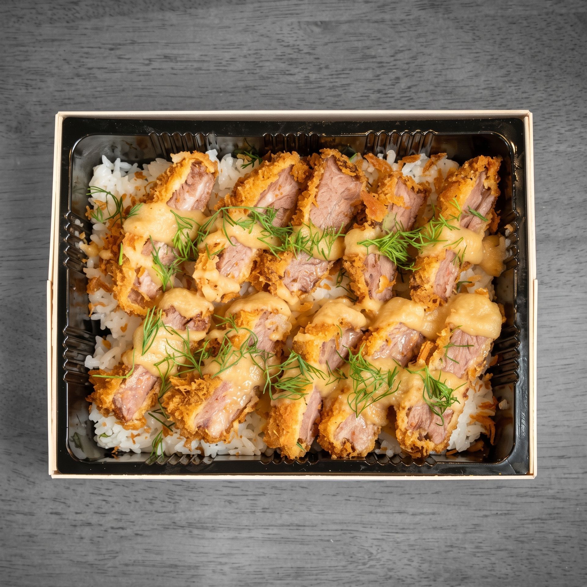 Top-down view of sushi rolls topped with sauce and herbs in a black tray on a gray table