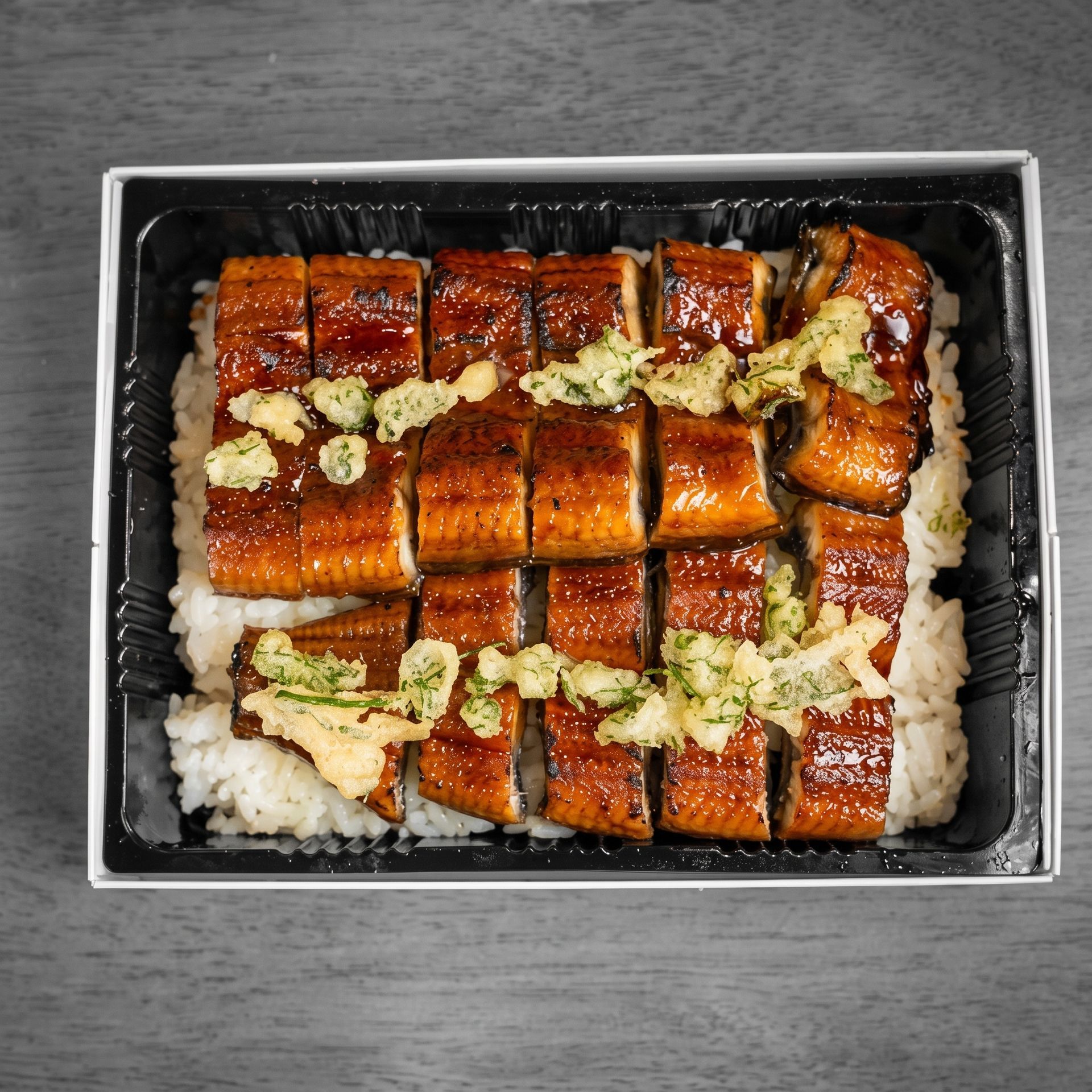 Takeout box of glazed grilled eel over white rice, topped with green sauce