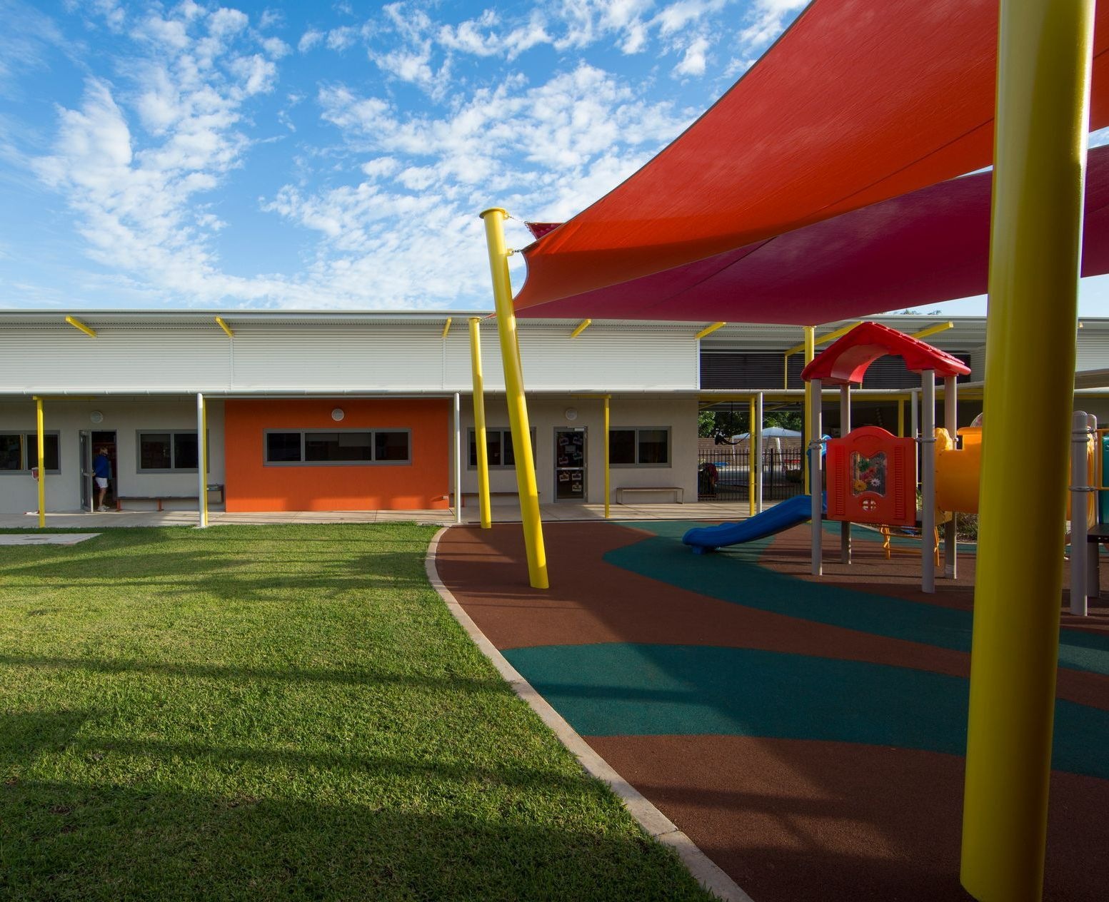A playground with a slide and a red umbrella in front of a building.