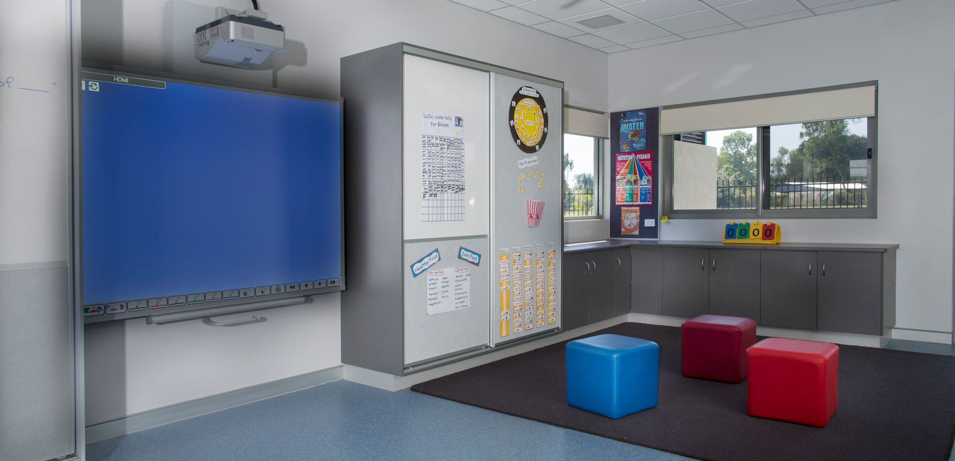 A classroom with a blue board , red ottomans , and a projector.