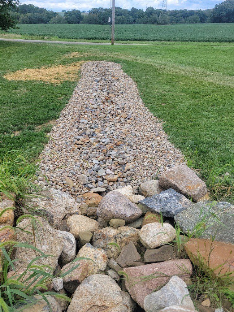 Gravel-lined waterway in a grassy field. Rocks border the bottom, leading to a long, raised gravel path.