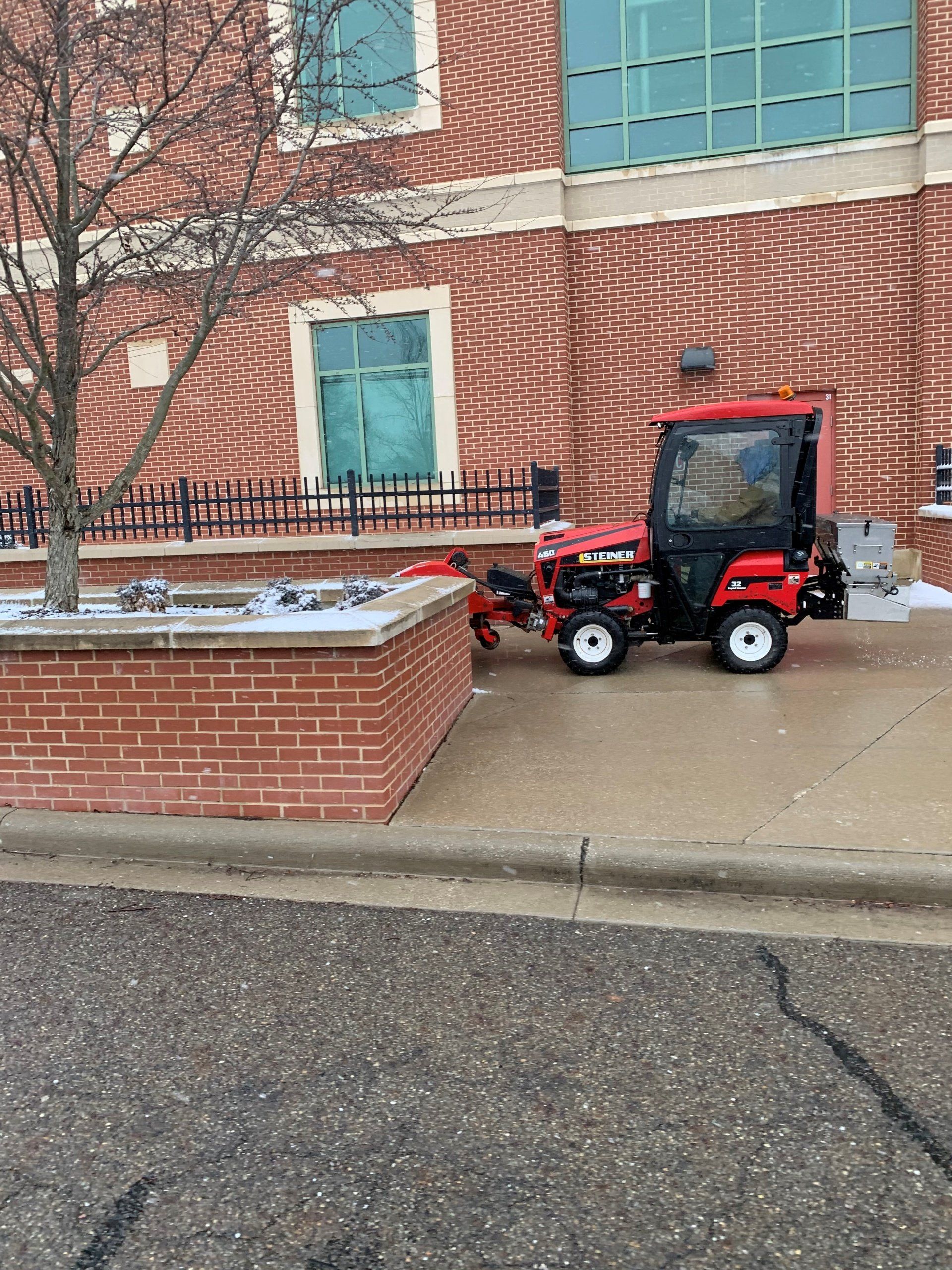 Red snow removal tractor on sidewalk next to a brick building and planter with snow.