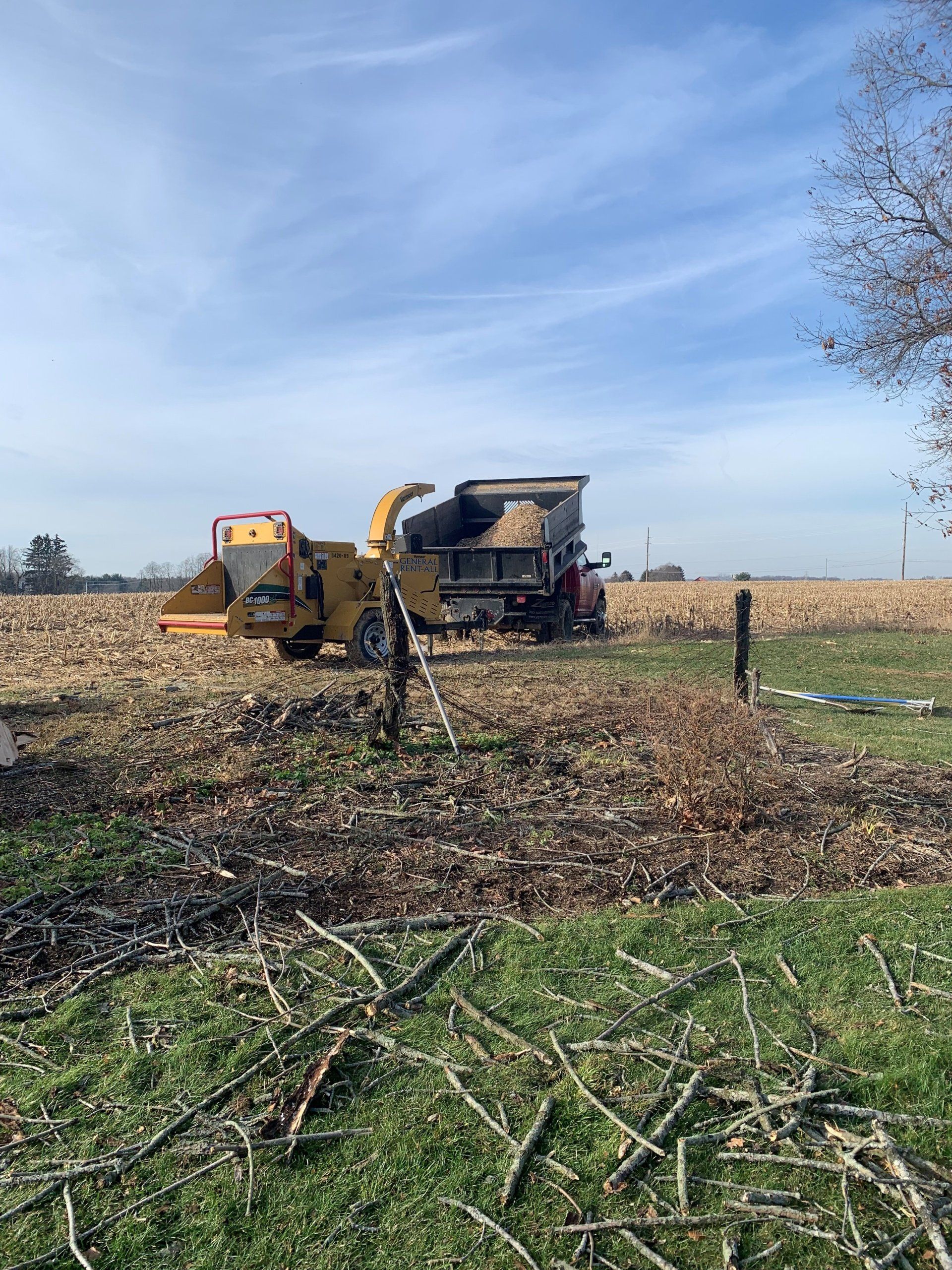 A wood chipper processes branches, loading a truck in a field, under a blue sky.