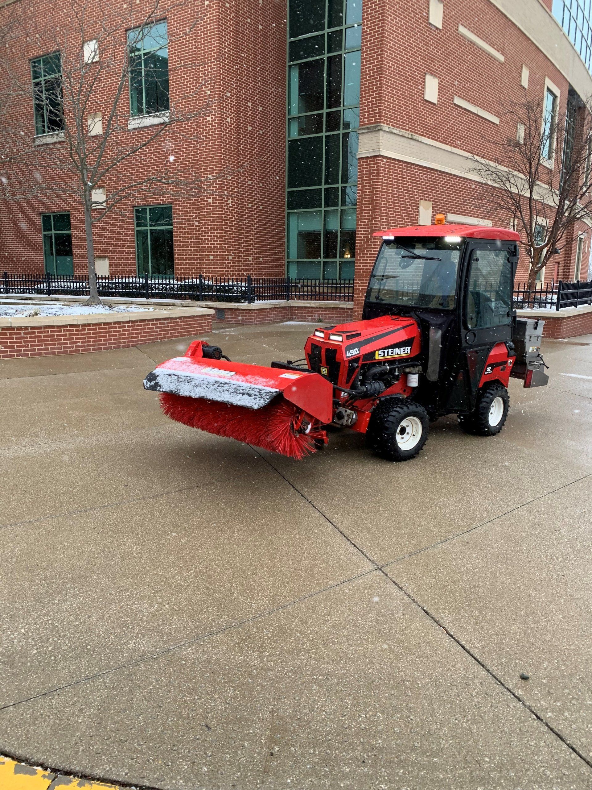 Red snow removal tractor with a brush on a brick building sidewalk.