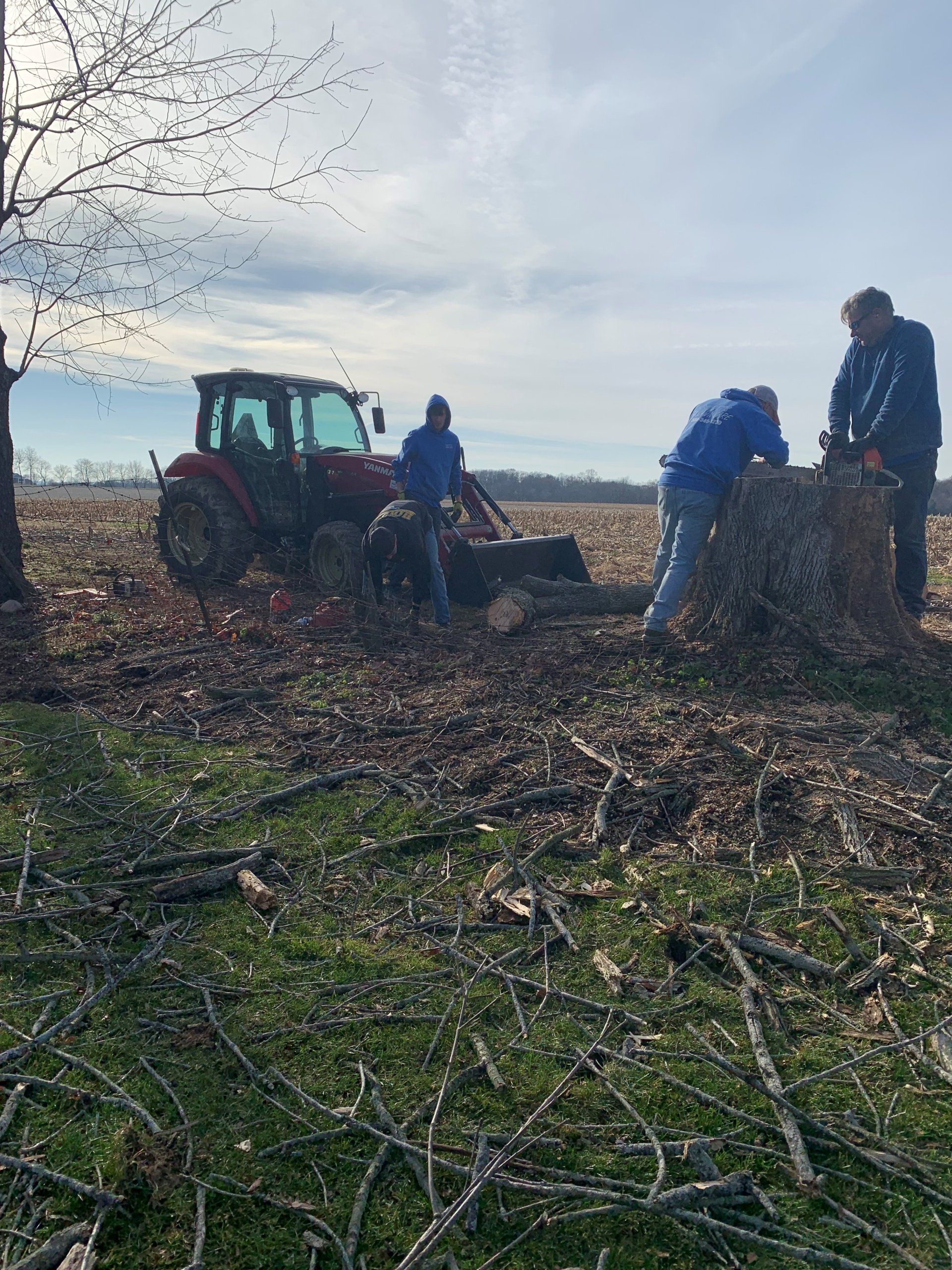 Three people using a tractor to clear brush and a large tree stump in a field.