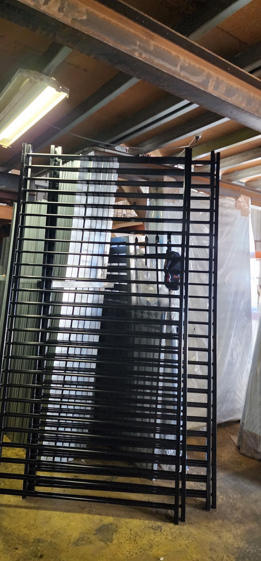 Black Metal Gates Stacked Inside a Warehouse, Under Exposed Beams — Metal Fencing Supplies In Taree, NSW