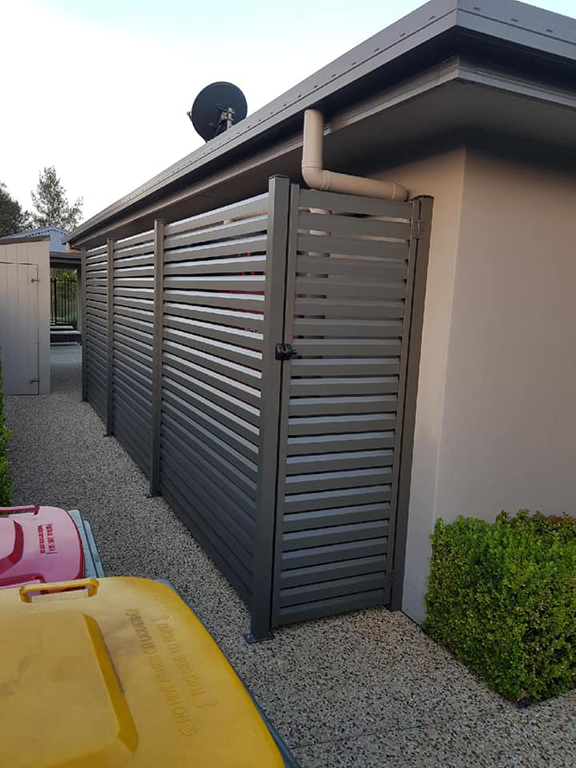 Gray Slatted Fence and Gate Alongside a Beige Building and Pebble Path — Metal Fencing Supplies In Taree, NSW