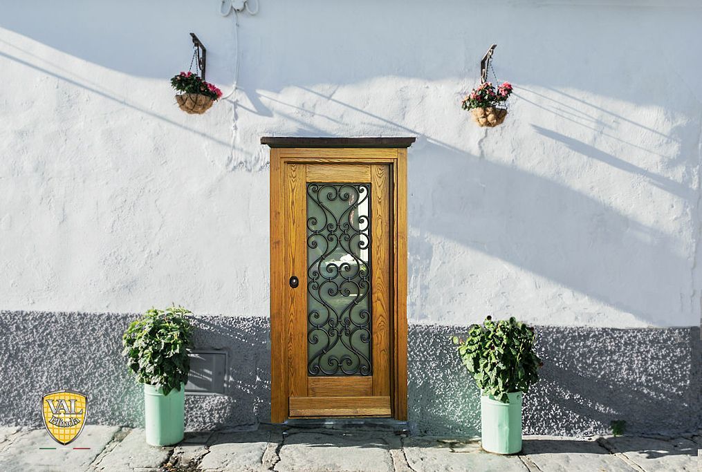 Porta d'ingresso in legno con vetri decorativi, fiancheggiata da piante in vaso su una parete intonacata di bianco.