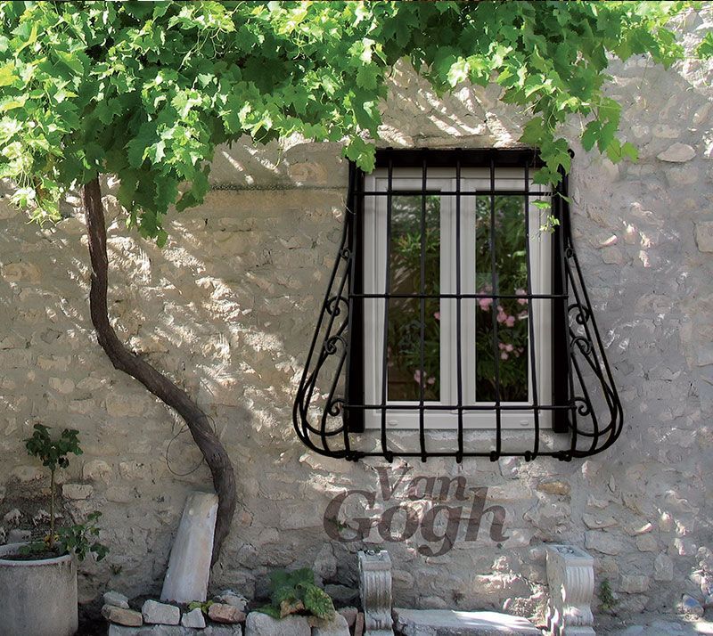 Muro in pietra con finestra sbarrata, albero frondoso e una piccola pianta in vaso in un cortile soleggiato.