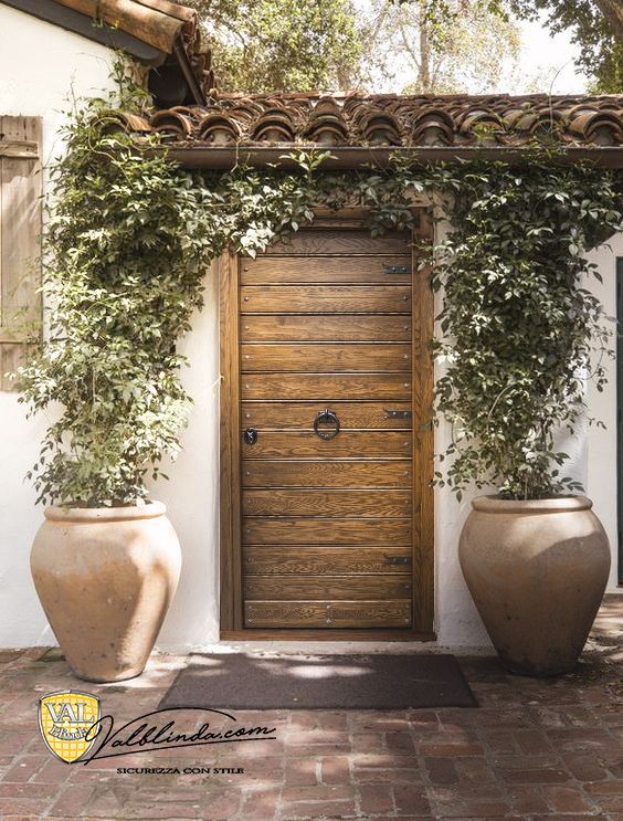 Porta d'ingresso in legno ricoperta di edera, fiancheggiata da due grandi urne di terracotta su un muro di stucco del cortile.