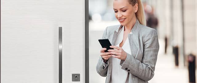 Una donna con un blazer grigio usa uno smartphone accanto a una porta bianca.