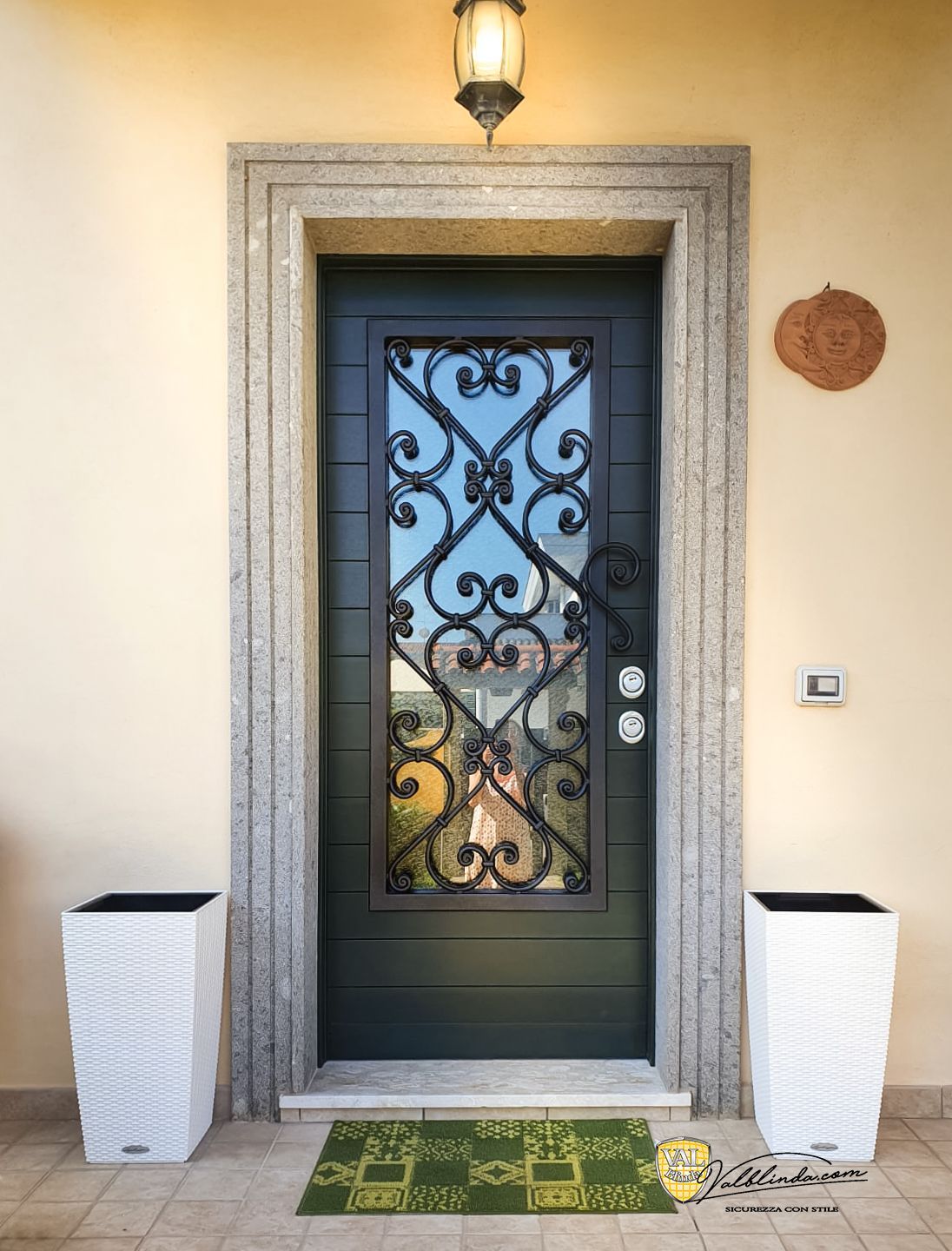 Ingresso con porta in vetro decorativo nero, cornici in pietra, lanterna e due fioriere bianche ai lati.