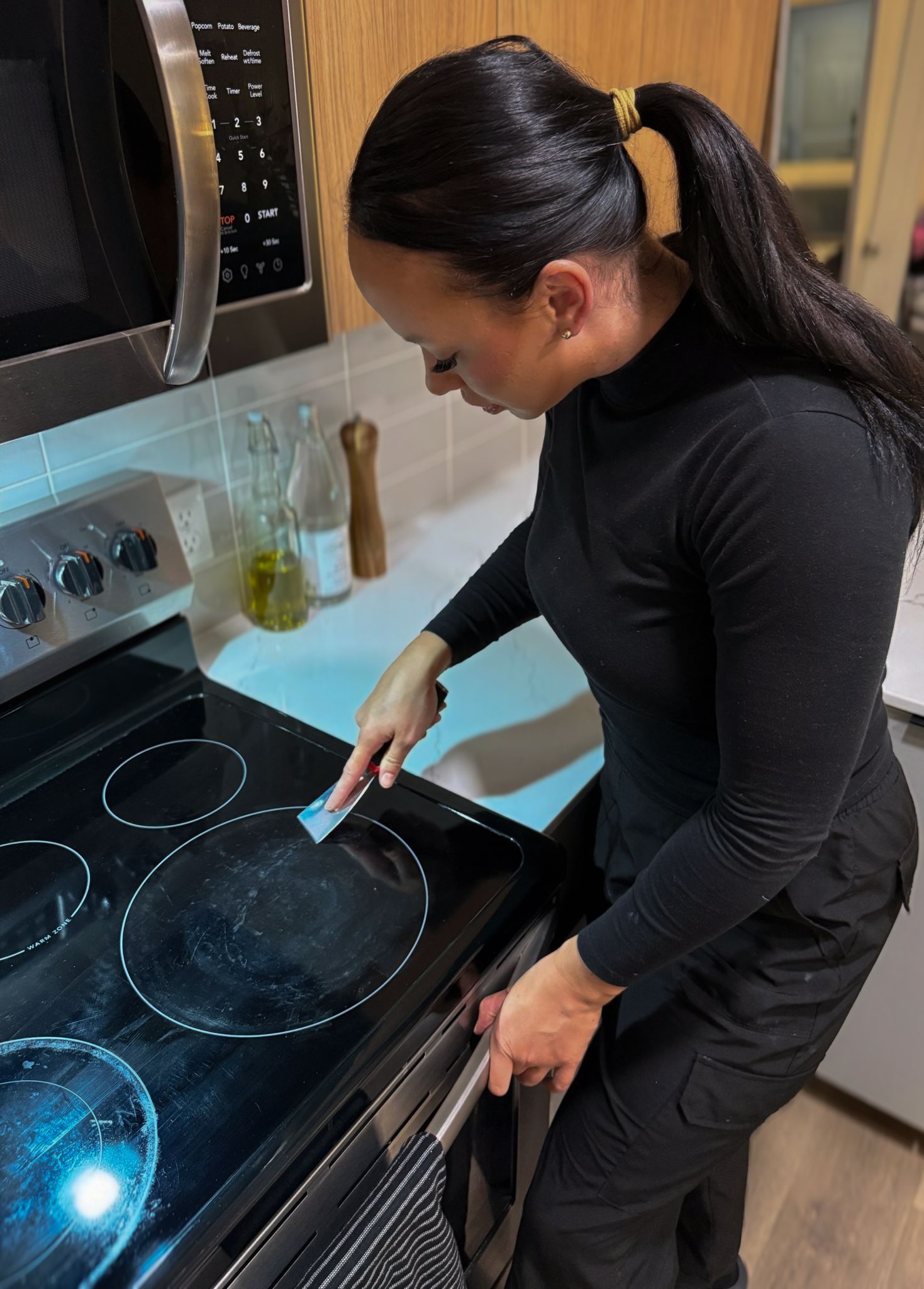 A person wearing a black long-sleeved shirt scrubs a glass stovetop with a small cleaning tool in a kitchen.