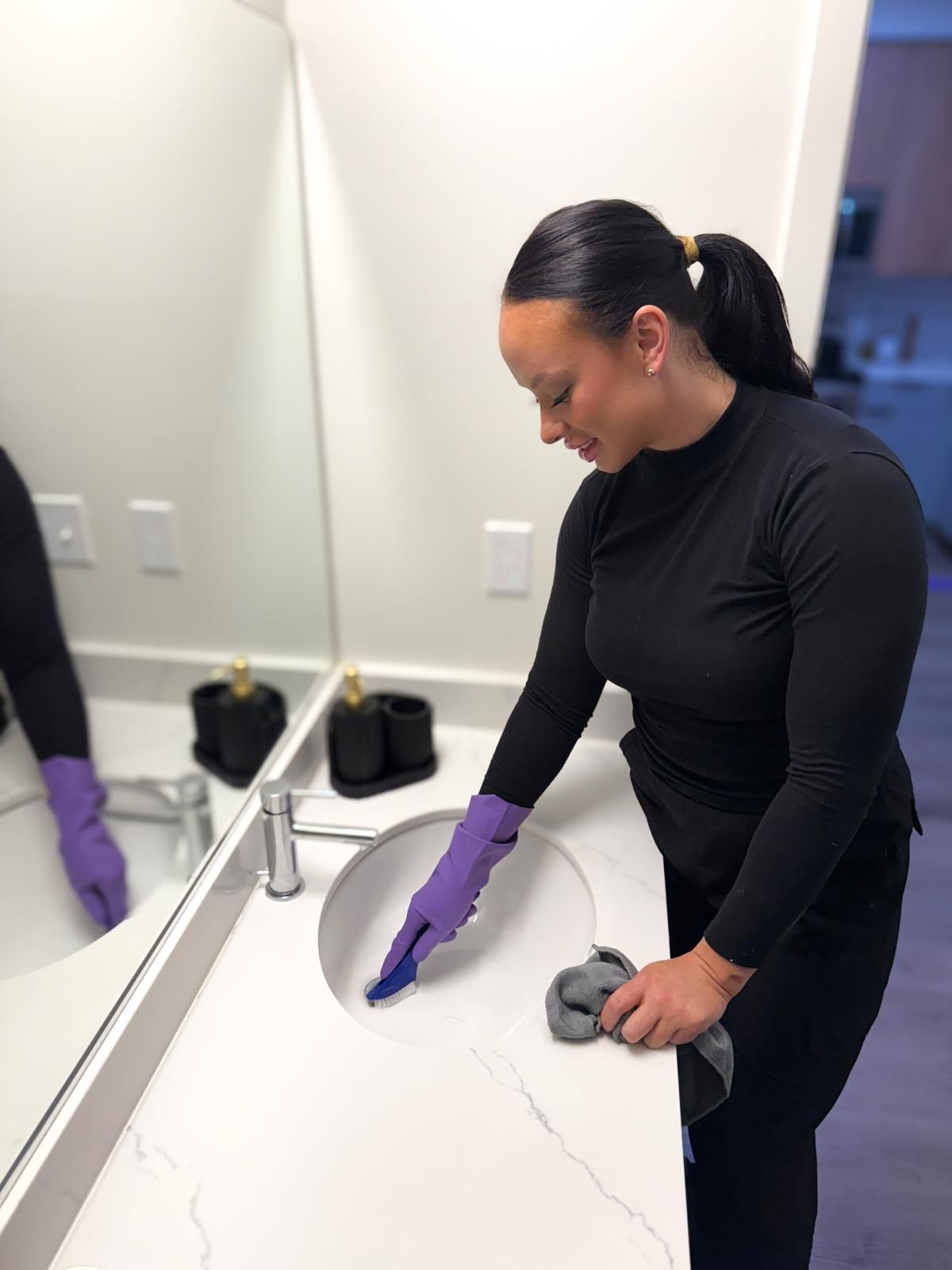 A person in a black shirt and purple gloves wipes a white bathroom sink with a grey cloth.