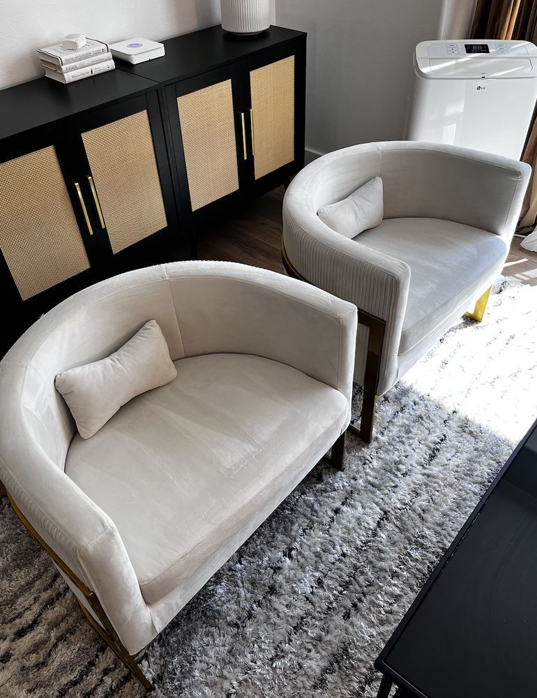 Two light-colored, curved velvet chairs sit on a grey patterned rug, in front of a black sideboard with rattan doors.