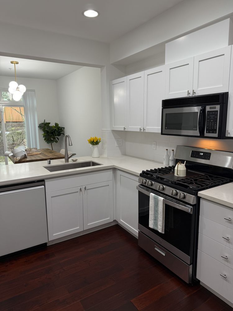 A modern kitchen with white cabinets, stainless steel appliances, dark wood floors, and a bright view into a dining area.