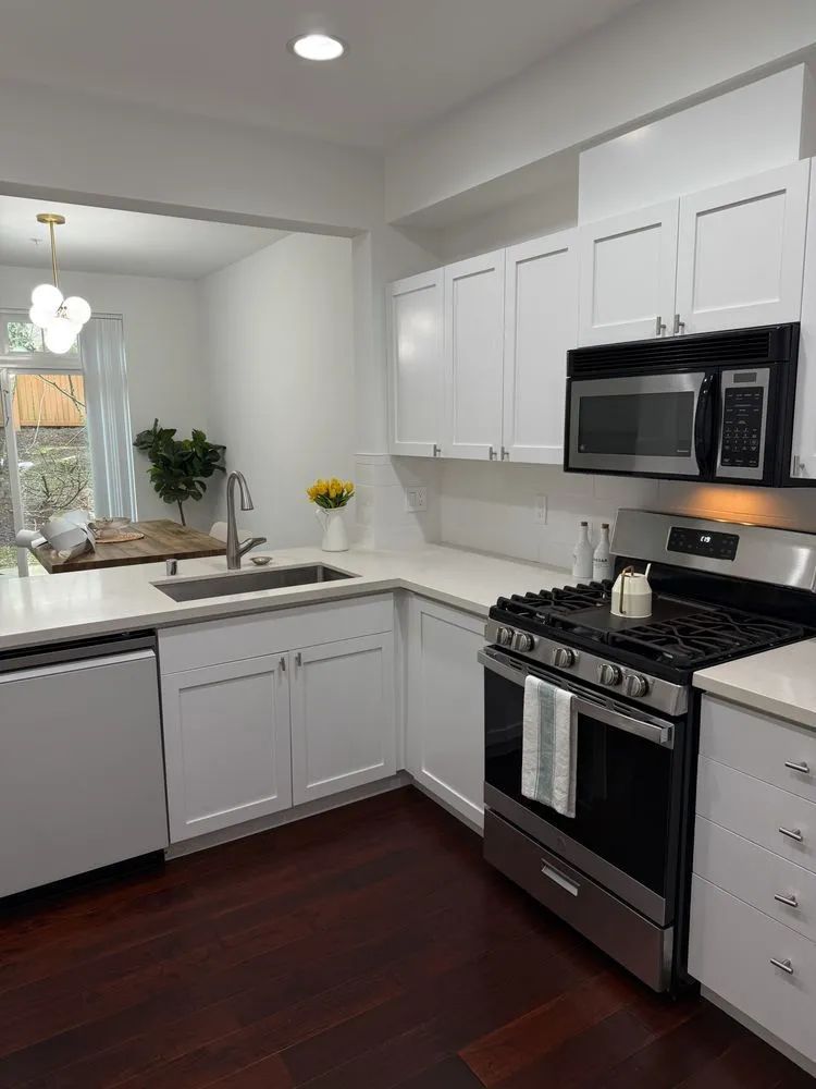 Bright white kitchen with black appliances, gray cabinets, dark wood floor, and sink by a window.