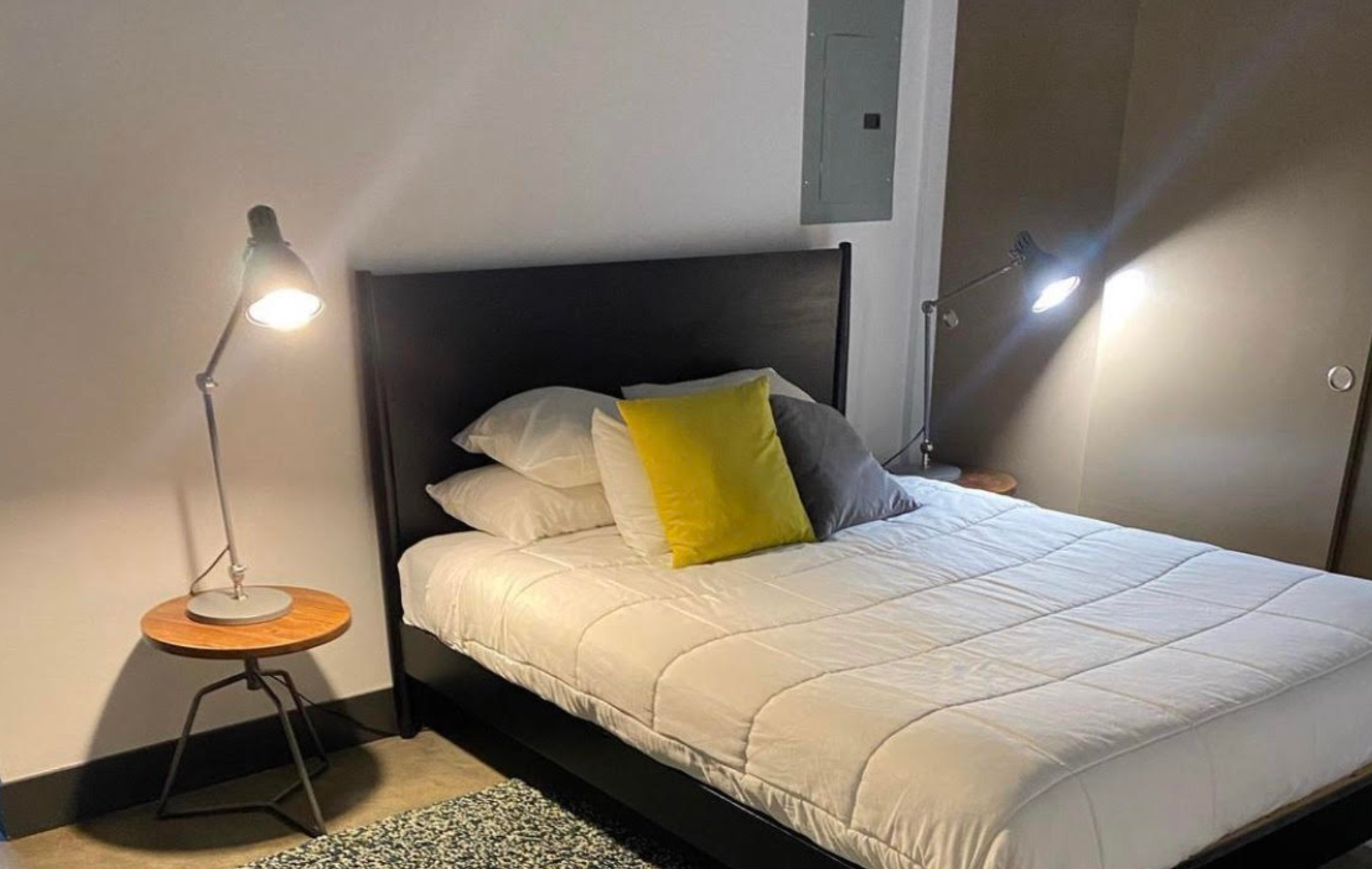 A bed with a dark headboard, white bedding, and yellow and gray accent pillows, flanked by two lamps on side tables.
