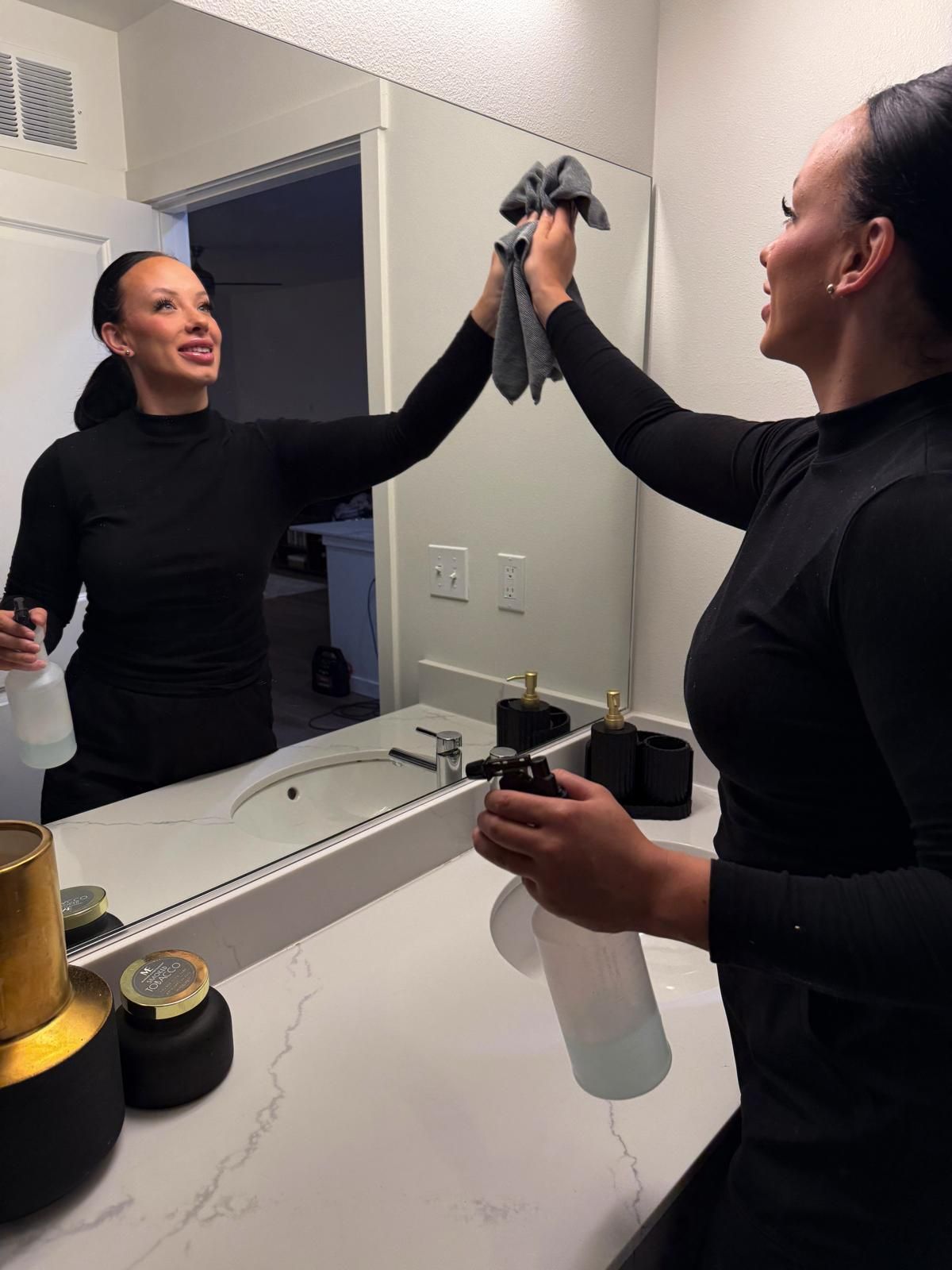 A person in a black long-sleeved shirt cleans a bathroom mirror with a cloth and a spray bottle.