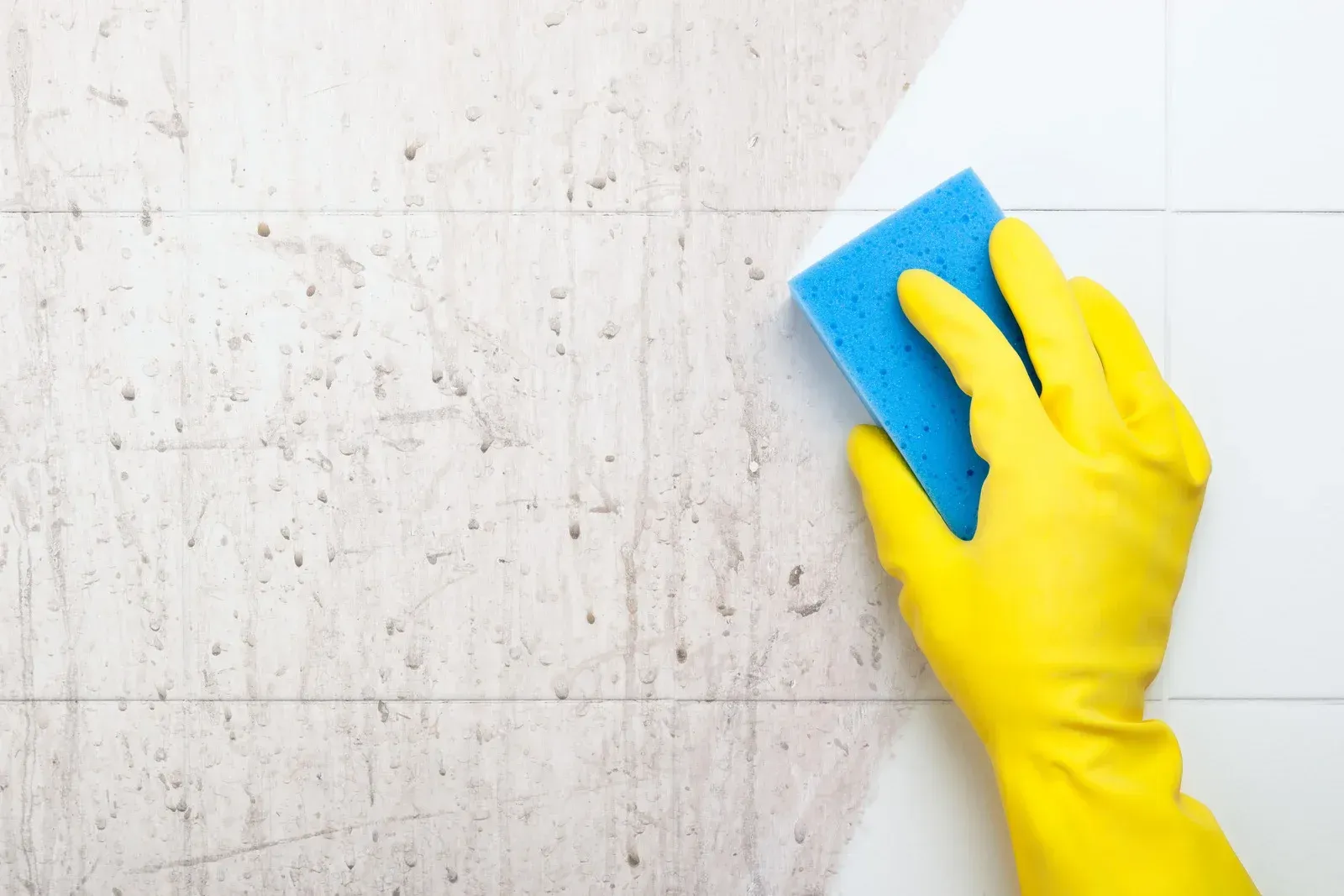 A person wearing yellow rubber gloves wipes a shelf with a blue cloth in a bright, modern room.