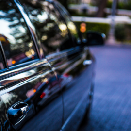 Close-up of a black car’s side door and window reflecting trees on a blurred street.