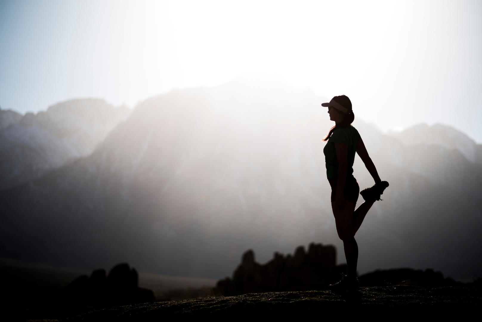 Silhouette of a woman stretching leg, mountain range in background.