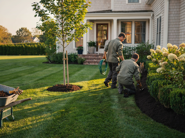 Two landscapers edging a lawn and garden beds beside a house at sunset
