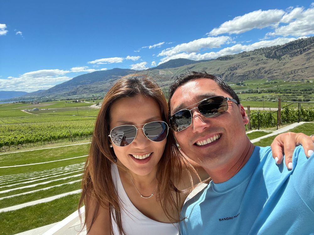 Justin Lee and his wife taking a selfie in sunglasses with green fields and mountains under a blue sky