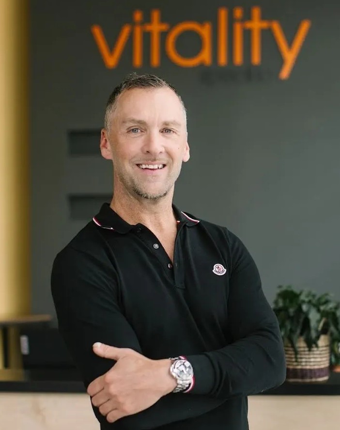 Smiling man in a black shirt standing indoors with an orange “vitality” sign behind him