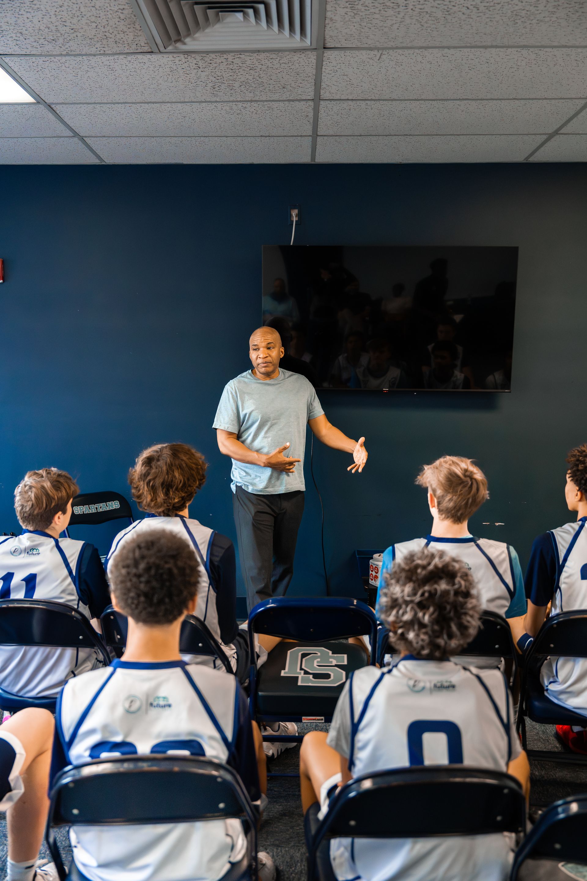 Coach speaking to team in a blue-walled room. Players wearing jerseys, seated, listening. TV on the wall.