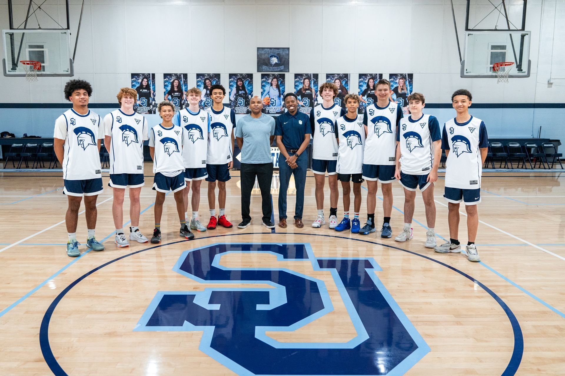 Basketball team poses on court with two coaches, wearing blue and white uniforms, in a gymnasium.