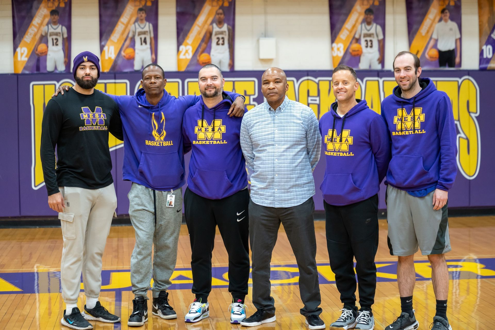 Six men in a basketball gym; coaches wearing purple hoodies, posing together. Banners in background.