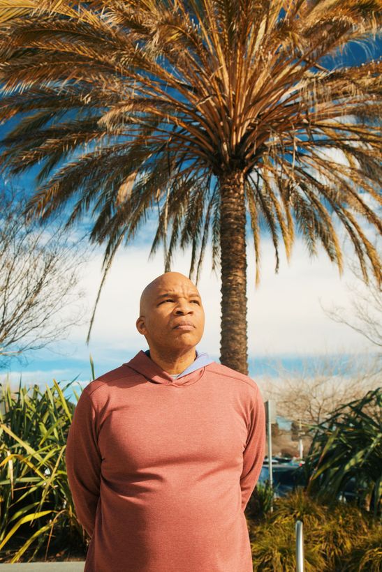 Man in a red hoodie stands under a palm tree, looking upward on a sunny day.