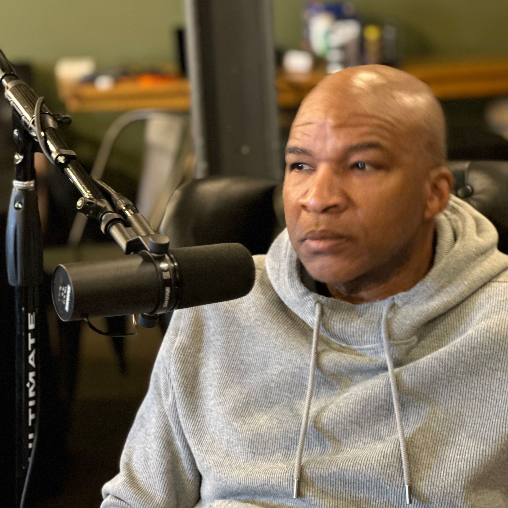 Bald Black man in gray hoodie sits by a microphone, looking off to the side.