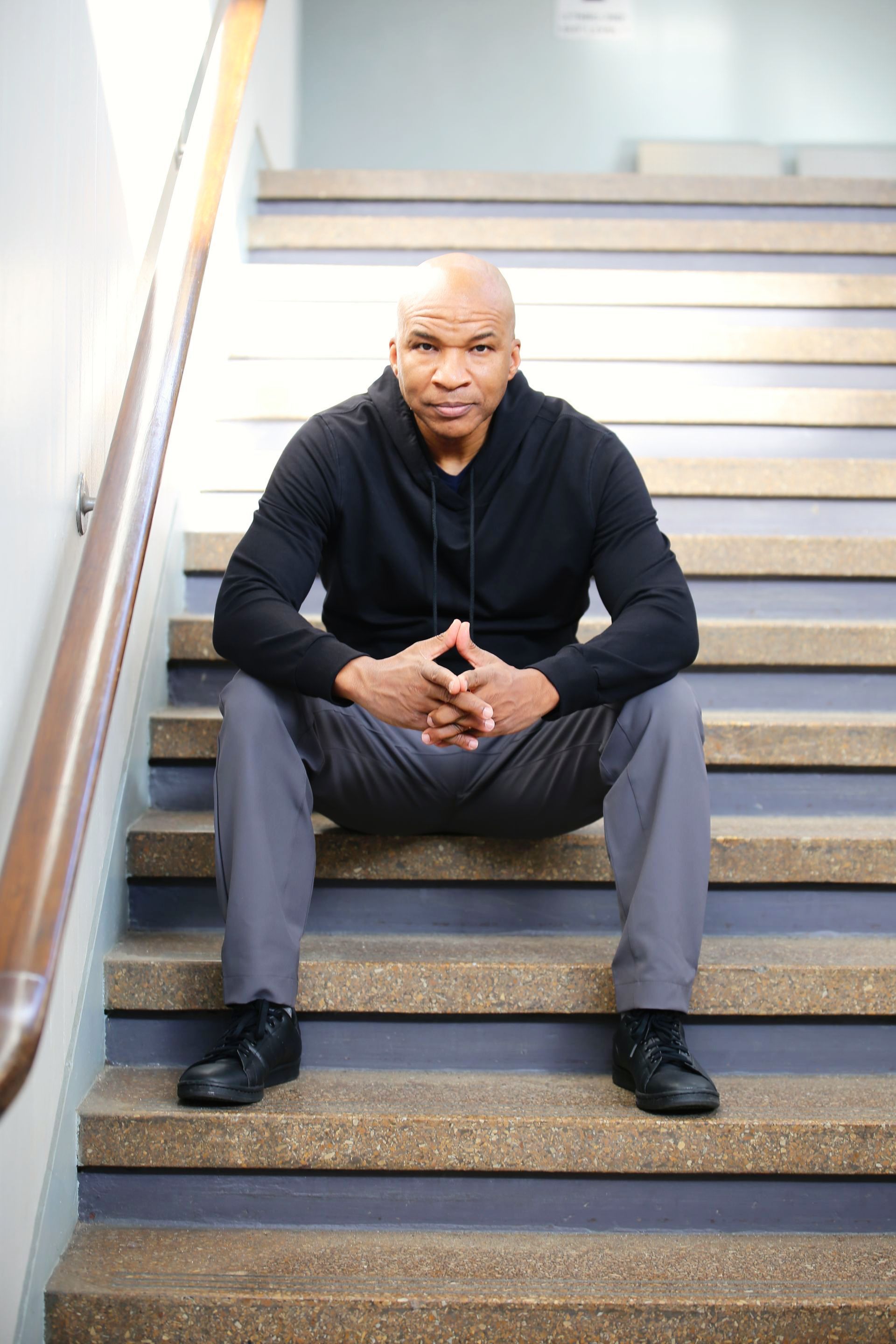 Man in black hoodie and gray pants sits on stairs, hands clasped, looking forward.