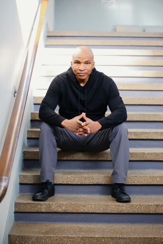Man in black hoodie and gray pants sits on stairs, hands clasped.