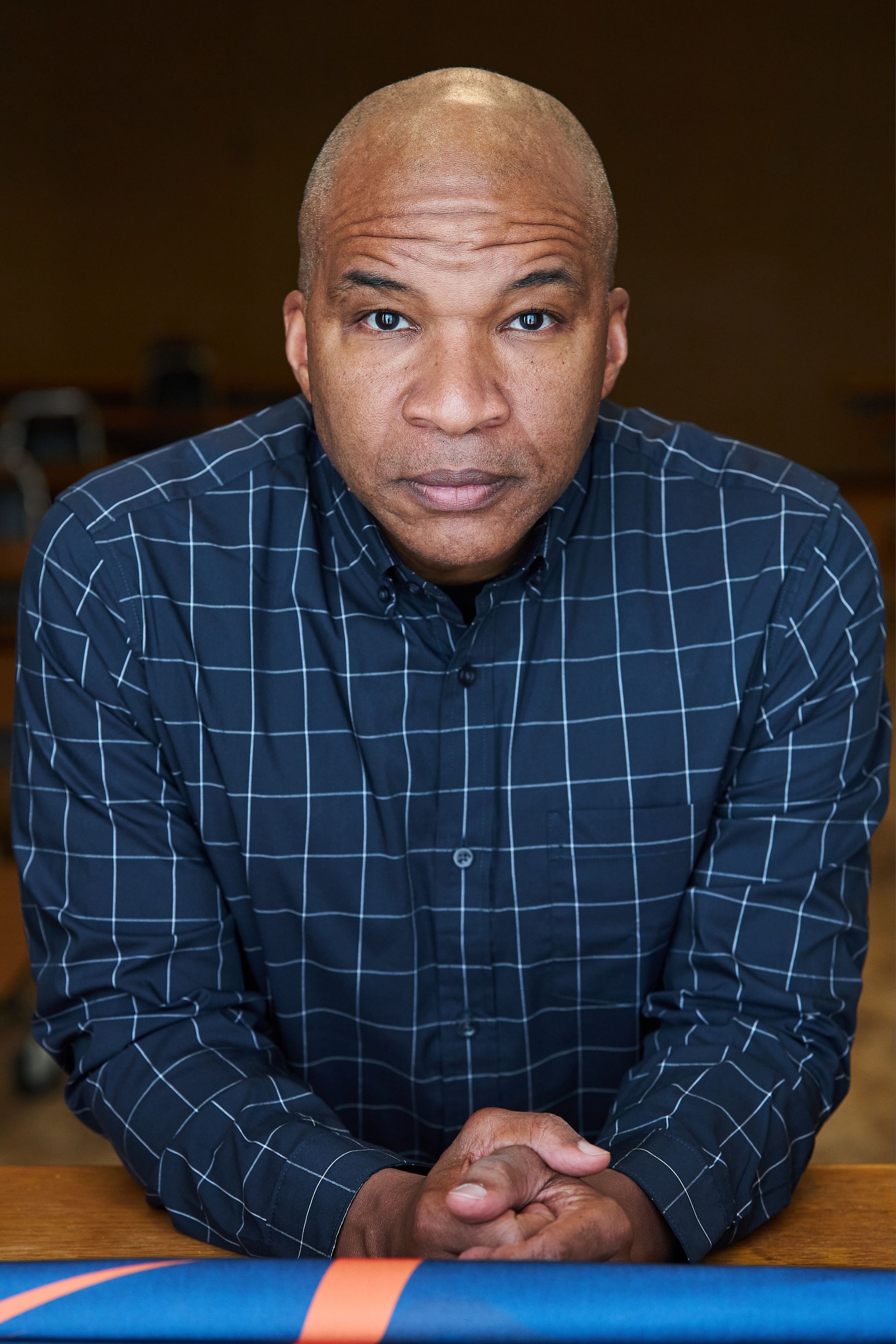 Bald Black man in blue plaid shirt, clasped hands, seated in a room, looking at the camera.